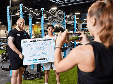 A gym member holding a whiteboard showing their new personal record while another person takes a photo with a smartphone