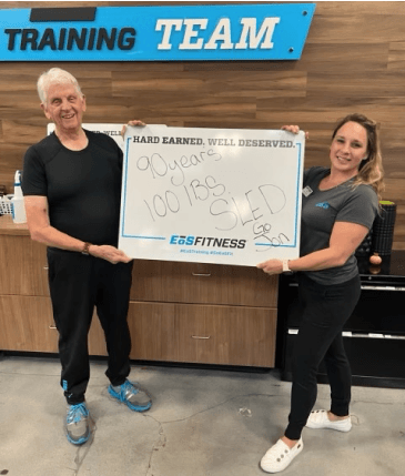 eos gym member standing side by side with personal trainer holding a white board listing how much weight they lifted