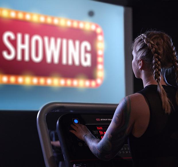 A person on treadmill is shown from the back, watching a movie screen.