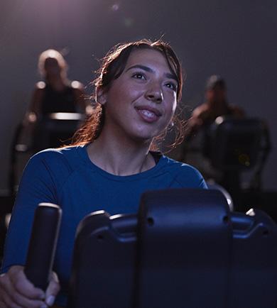 Two women are exercising on stationary bikes, smiling and engaged in their workout.